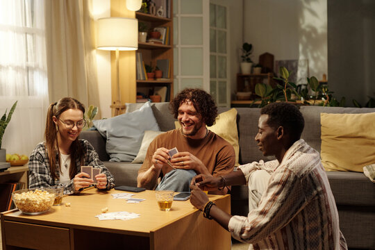 Happy intercultural friends playing cards at leisure while gathered around table in spacious living room