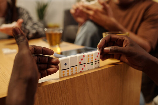 Hands of young African American man sitting by table with row of domino tiles during tabletop game