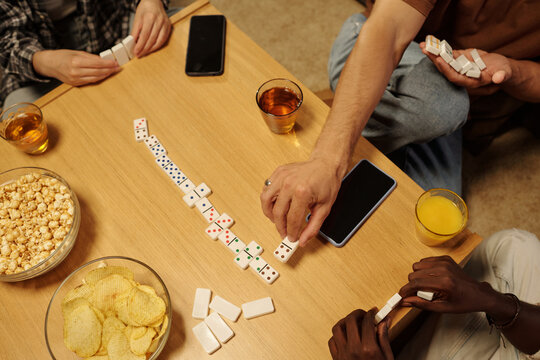 High angle of hands of three young intercultural friends playing domino by wooden table at leisure