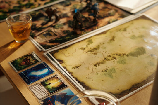 Maps and pictures for tabletop game lying on table next to row of cards with color illustrations and glass of juice