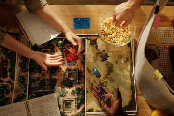 Top view of hands of three young intercultural friends playing tabletop game by table and having popcorn
