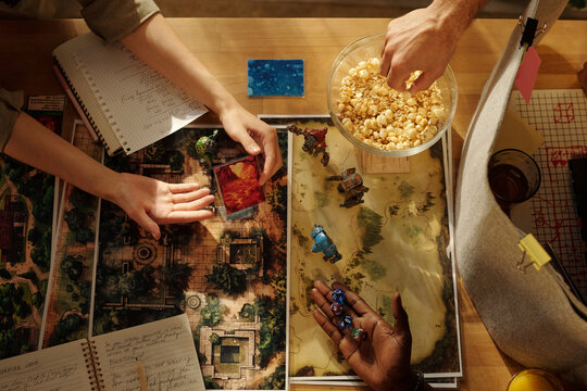 Top view of hands of three young intercultural friends playing tabletop game by table and having popcorn