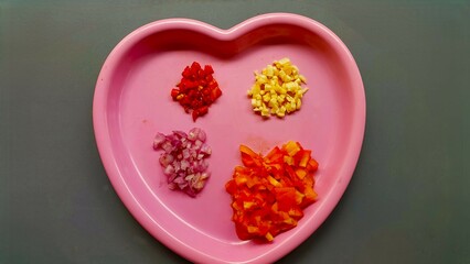A pink heart-shaped plate containing four types of chopped ingredients: red onion, red chili, garlic, and tomato. It is interesting because it shows the preparation of the ingredients.