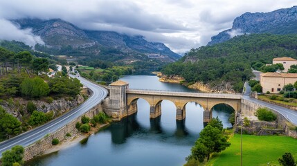 Fototapeta premium Scenic view of an ancient bridge spanning a serene river surrounded by mountains in a tranquil landscape
