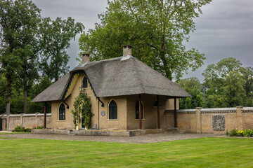 Fototapeta premium A charming brick house with a thatched roof, arched windows, and a green door covered in climbing plants. The house sits in a lush garden, with a cobblestone path leading to the entrance.