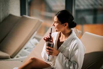 Woman enjoying a relaxing spa day in a white robe with a refreshing cocktail in hand during a...