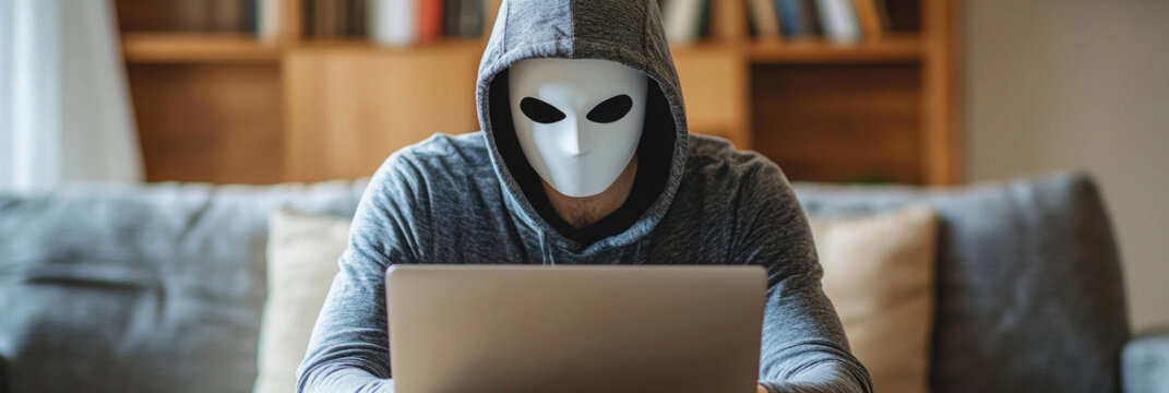 Anonymous figure focused on laptop with a mask sitting in a cozy living room