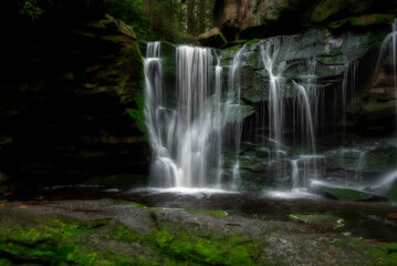 A serene waterfall flows gracefully over moss-covered rocks, surrounded by lush greenery.