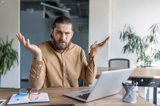 A man with a puzzled expression sits at a desk using a laptop, gesturing with raised arms, conveying confusion or hesitation while working in an office or professional setting.