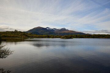 Naklejka premium A peaceful view of Lough Leane with its calm waters reflecting the surrounding landscape and distant mountains, located near Ross Castle in County Kerry, Ireland. An idyllic natural scene.