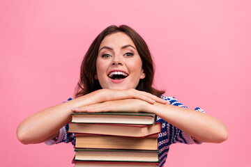 Cheerful young woman with brunette hair leaning on stack of books against pink background, exuding...