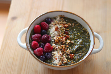 Beige bowl with green smoothie, frozen berries and homemade granola, served on the table. Selective focus.