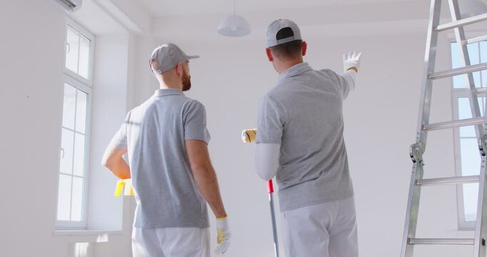 Back view of two painters in work attire standing in bright room and discussing apartment renovation plan. Professional male contractor workers having conversation about design project at new house.