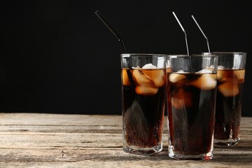 Refreshing cola with ice cubes and drinking straws in glasses on wooden table against black background, closeup. Space for text