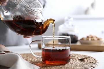 Woman pouring hot black tea into cup at white table, closeup