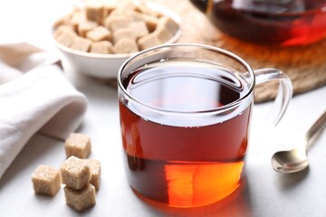 Aromatic black tea in cup and brown sugar cubes on white table, closeup
