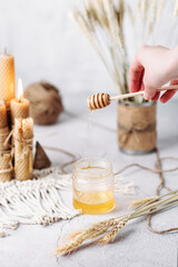 A woman holds a wooden honey spoon in her hands, from which liquid spring honey flows, a dawn composition, wax candles in the background, wheat ears, macrame, honey breakfast