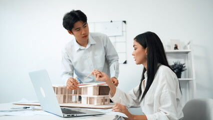 Professional male asian architect using ruler to measure house model length while young beautiful caucasian colleague using laptop to analyzed data on meeting table with house model. Immaculate.