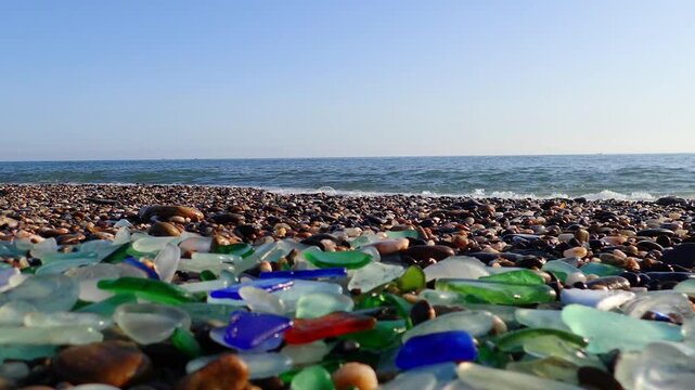 Colored sea glass with beach pebbles and shells in the mediterranean coast and in the background sea and waves with sunset and sky, sandy beach with coast glass, beach glass.