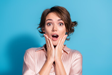 Young woman with surprised expression holding face on blue background wearing pink satin blouse