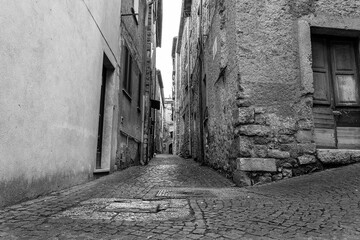 Obraz premium Black-and-white photo of a narrow alley with small stone pavement with manhole covers, flanked by old medieval stone houses with wooden gates
