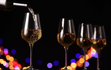 Pouring white wine into glass against black background with blurred lights, closeup. Bokeh effect