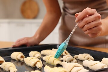Woman brushing egg wash onto raw croissants at table indoors, closeup