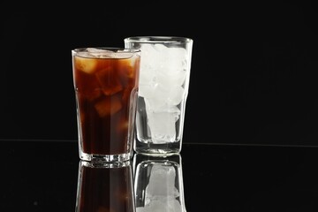 Refreshing iced coffee and ice cubes in glasses on black mirror surface. Space for text