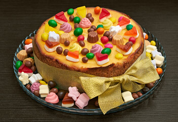 Festive cheesecake decorated with colorful candies, jelly, and chocolate, placed on a glass platter with a golden bow.