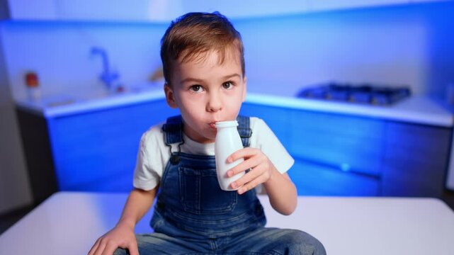 Brunet Caucasian toddler boy wearing white t-shirt and jeans romper drinks yogurt from a little bottle. Kid sitting on the table having healthy snack. Close up.