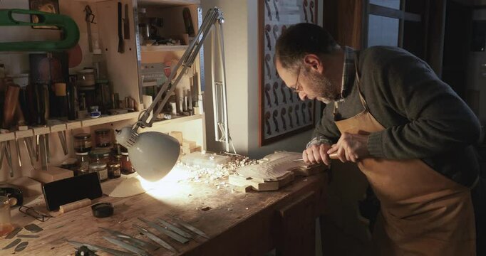 Slow motion shot of wood chips and shavings flying out as caucasian maker is carving belly bottom plate for a violin using gouge in workbench, tools in background, crafting a musical masterpiece