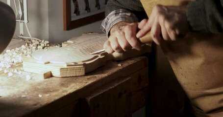 Luthier working on top plate of a violin using a gouge, with wood shavings on workbench and tools in background, crafting a musical masterpiece with precision and expertise close up shot