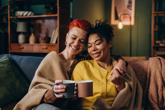 Lesbian couple holds tea cups sharing gentle hug sitting on sofa in evening
