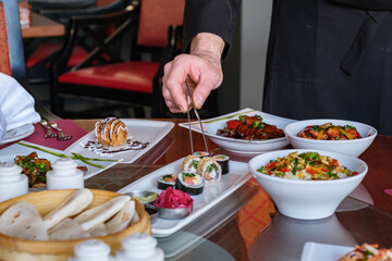 A chef’s hand using tongs to carefully place sushi rolls on a plate at Asian culinary restaurant.