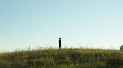 Fototapeta premium Solitary figure standing on grassy hill under clear sky.