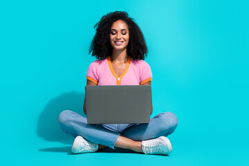 Naklejka premium Young woman sitting cross-legged, using laptop on turquoise background, wearing pink top and jeans, smiling and relaxed.
