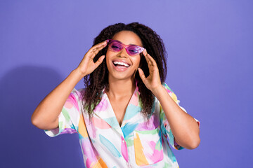 Vibrant young woman with curly hair in trendy outfit and purple glasses smiles happily against a violet background