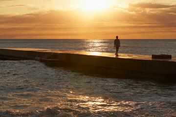 A lone figure of a man on a pier in the light of the setting sun. The sea surface is colored with golden light by the setting sun. Silence and solitude. Copy space.