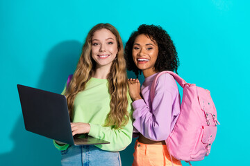 Two cheerful young women in casual sweatshirts with laptop and backpack in front of a turquoise...