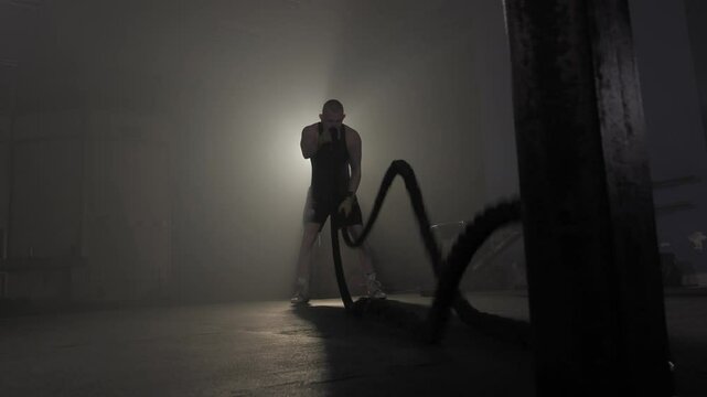 Boxer training with battle ropes in a smoky, atmospheric gym setting.