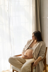 Stylish beautiful pregnant woman in beige suit holding baby bump and posing on chair in soft light. Fertility, health care and expecting new life concept