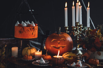 Beautiful table with homemade delicious cake for Halloween party with meringue ghosts and chocolate pumpkins. Candles in chandelier, spider web, jack-o-lantern for festive holiday decoration. Dark