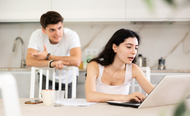 Young guy screams during family quarrel with young woman using laptop in kitchen