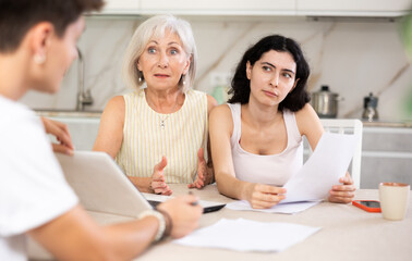 Young girl with elderly retired mother is talking to guy who is real estate sales manager. Man talks about advantages of residential complex and credit conditions when buying home
