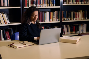 Woman studying in library using laptop with open books on the table