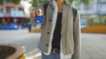 Young man holding credit card in urban park setting showing modern financial lifestyle with focus on hand and city elements in vibrant outdoor environment