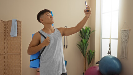 Young asian man taking a selfie in a wellness spa room with yoga mat and fitness items, wearing a...