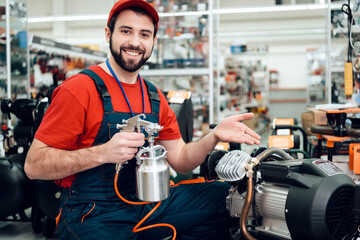 Salesman in red shirt and baseball cap is posing with compressor paint sprayer on foreground in...
