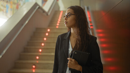 Young, hispanic, woman in a lobby with stairs, holding tablet, wearing glasses and blazer, looking sideways, modern, interior, illuminated by red lights on stairs