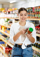 Interested smiling young girl scanning barcode on glass jar of canned food with smartphone while shopping in supermarket, paying for groceries using mobile app
